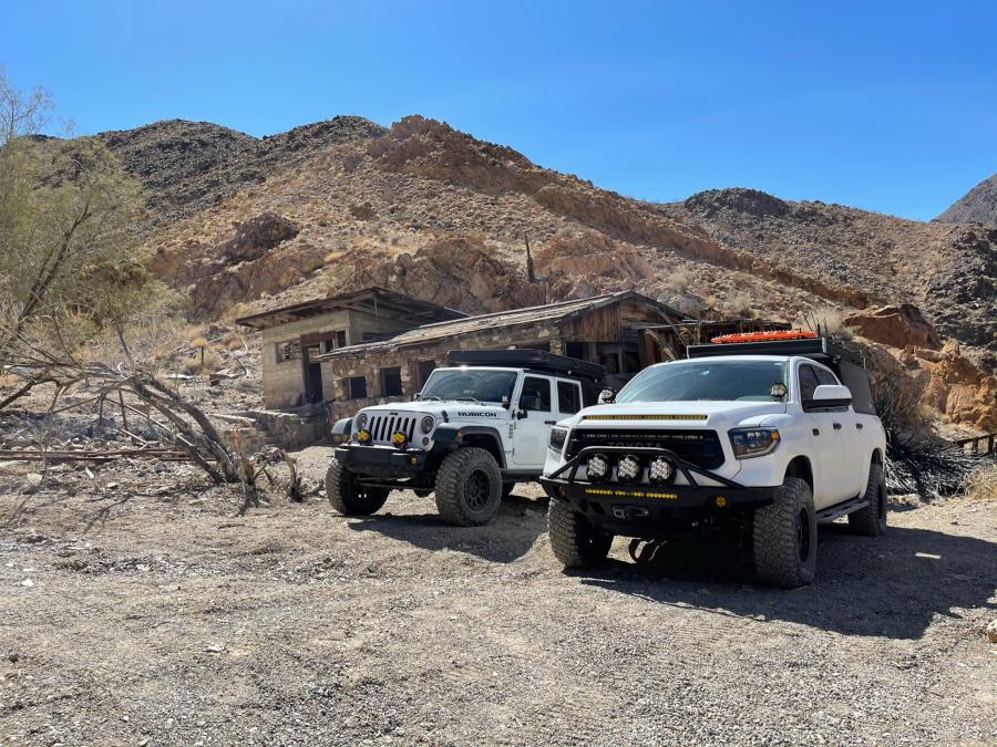 White Jeep Wrangler and Toyota Tundra parked beside a crumbling stone-and-wood mining cabin at the base of desert mountains.