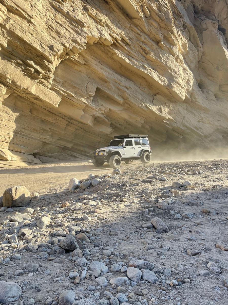 White Jeep Wrangler with a rooftop tent kicking up dust while driving through a narrow slot canyon with striated sandstone walls.