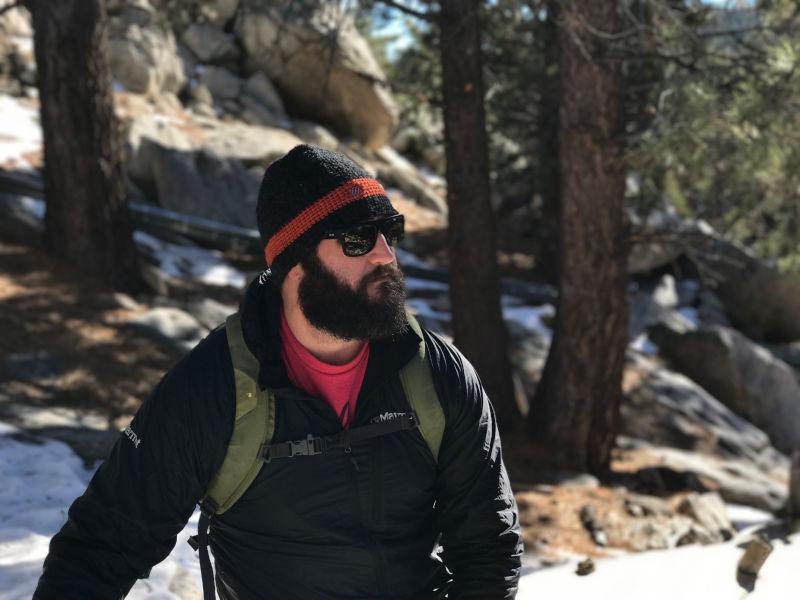 Hiker in a black jacket and orange-striped beanie pausing on a snowy pine forest trail with granite boulders.