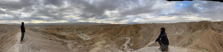 Two hikers standing on a desert ridge overlooking badlands terrain under a cloudy sky.