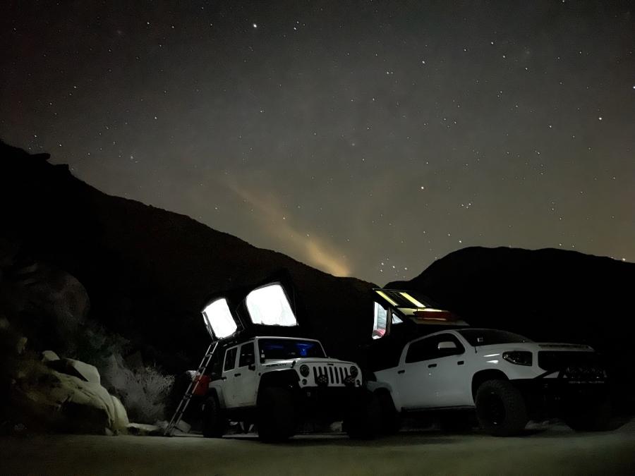 Two overlanding rigs with rooftop tents glowing at night, parked in a desert canyon under a star-filled sky.