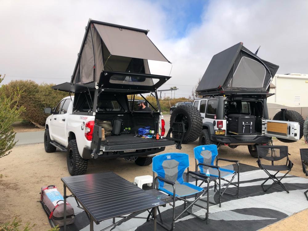 Jeep Wrangler and Toyota Tundra with rooftop tents open at a campsite, camp chairs and a table set out for the evening.