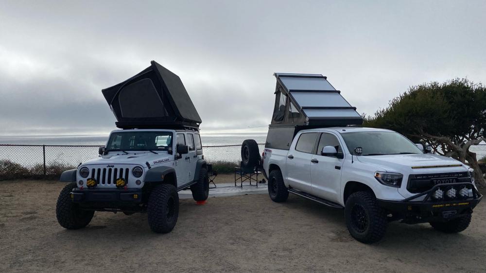 Jeep Wrangler and Toyota Tundra with rooftop tents open at a bluff-top campsite overlooking the fog-covered ocean.
