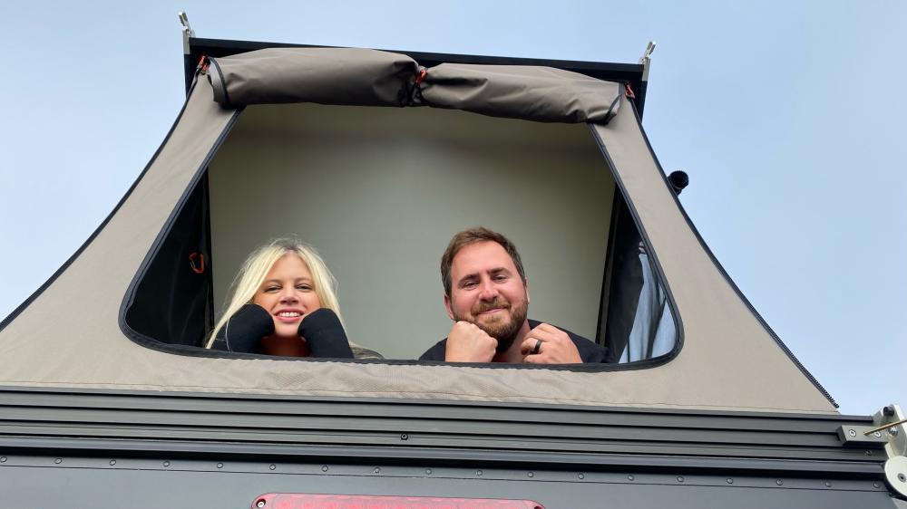 Couple smiling out through the open rooftop tent window against a clear blue sky.