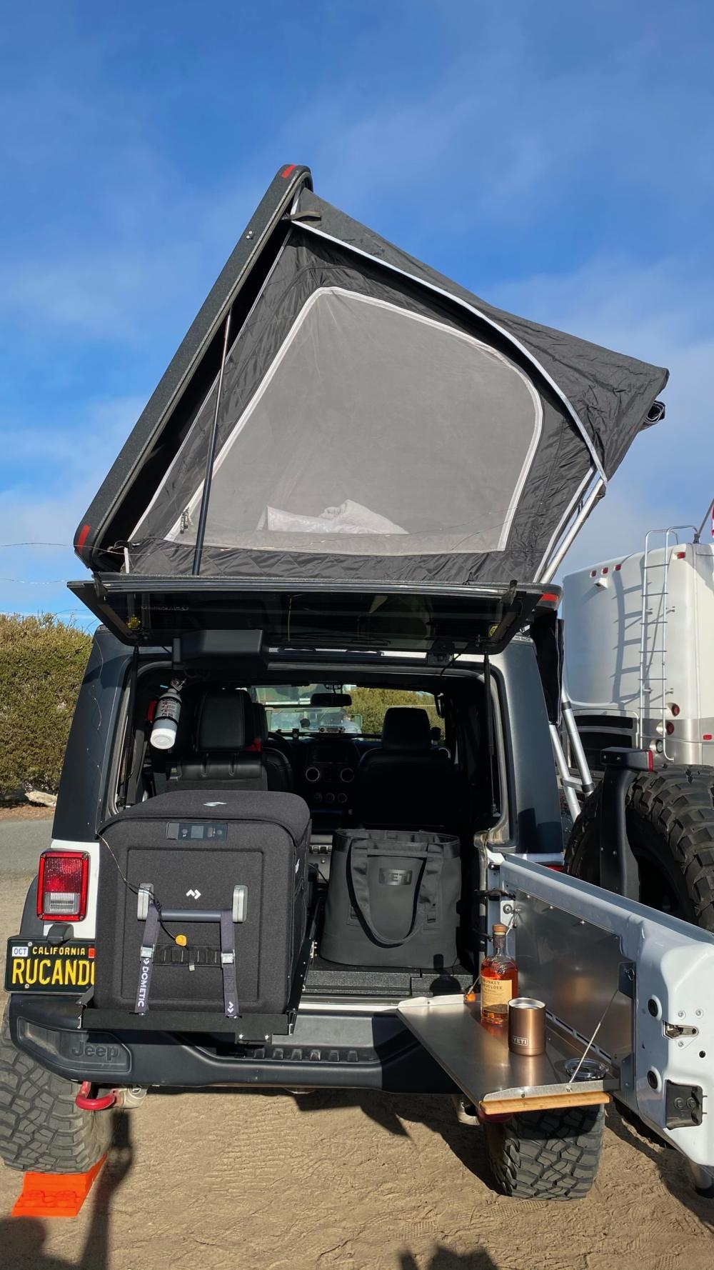 Open rooftop tent on a Jeep Wrangler with bedding visible inside, tailgate loaded with gear and a bottle of whiskey set out on a fold-down table.