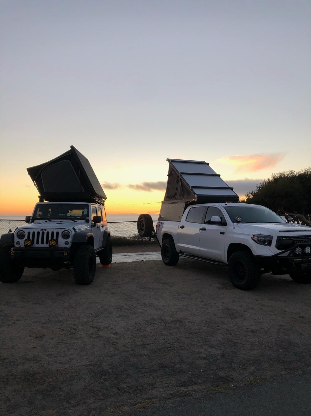 Jeep Wrangler and Toyota Tundra with rooftop tents open, parked by the ocean as the sun sets in orange and pink.