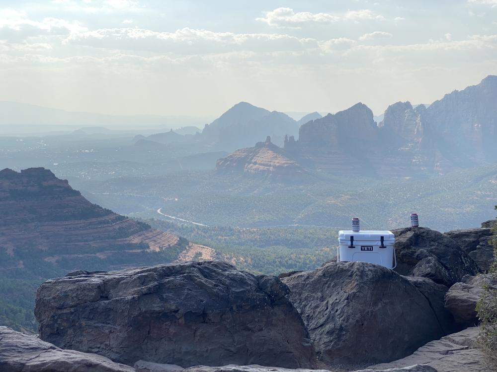 YETI cooler with two beer cans balanced on a cliff edge overlooking Sedona's red rock canyons and buttes in hazy afternoon light.