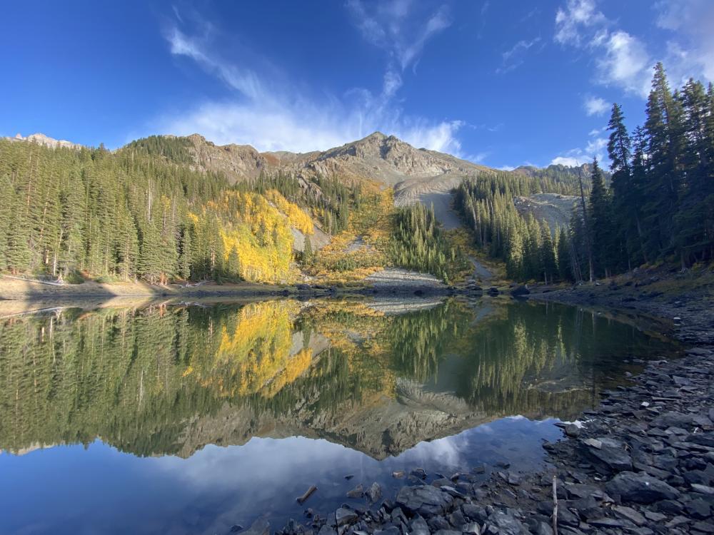 Alpine lake near Ophir mirroring pine forest, golden aspens, and rocky peaks under a blue sky.