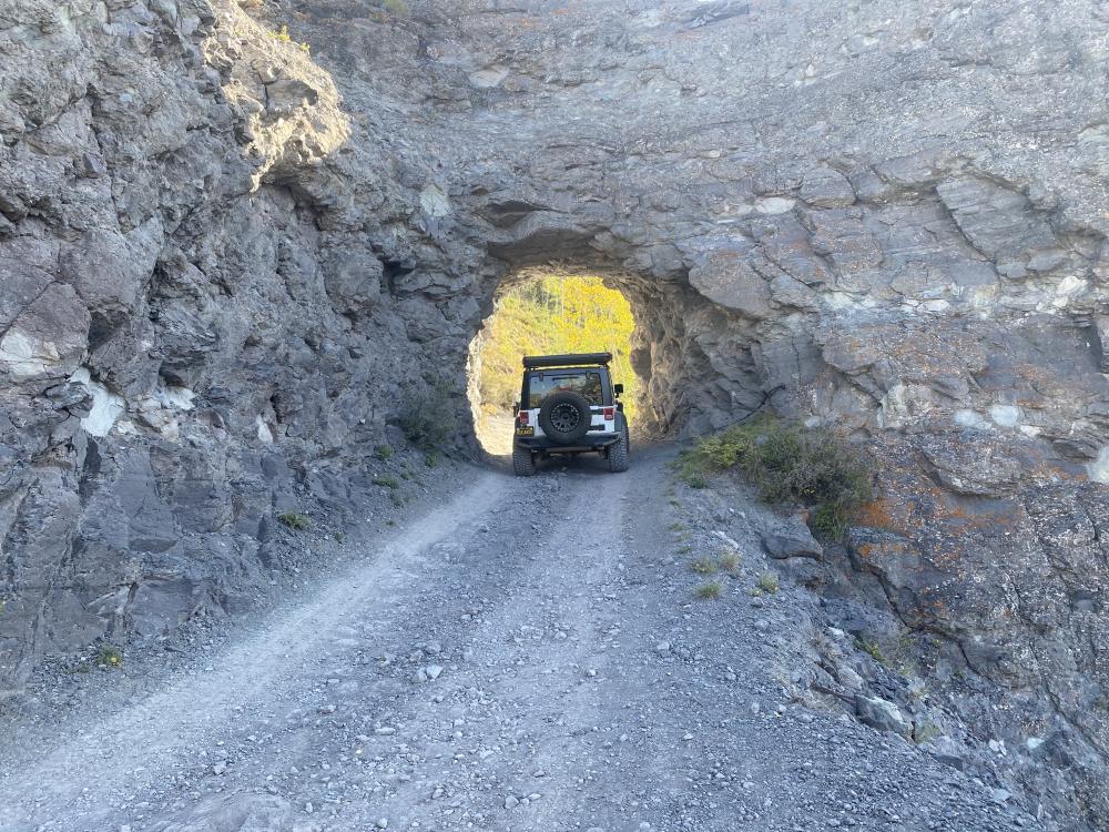 White Jeep Wrangler driving through a rock tunnel blasted through the mountainside outside Telluride, golden aspens visible beyond.