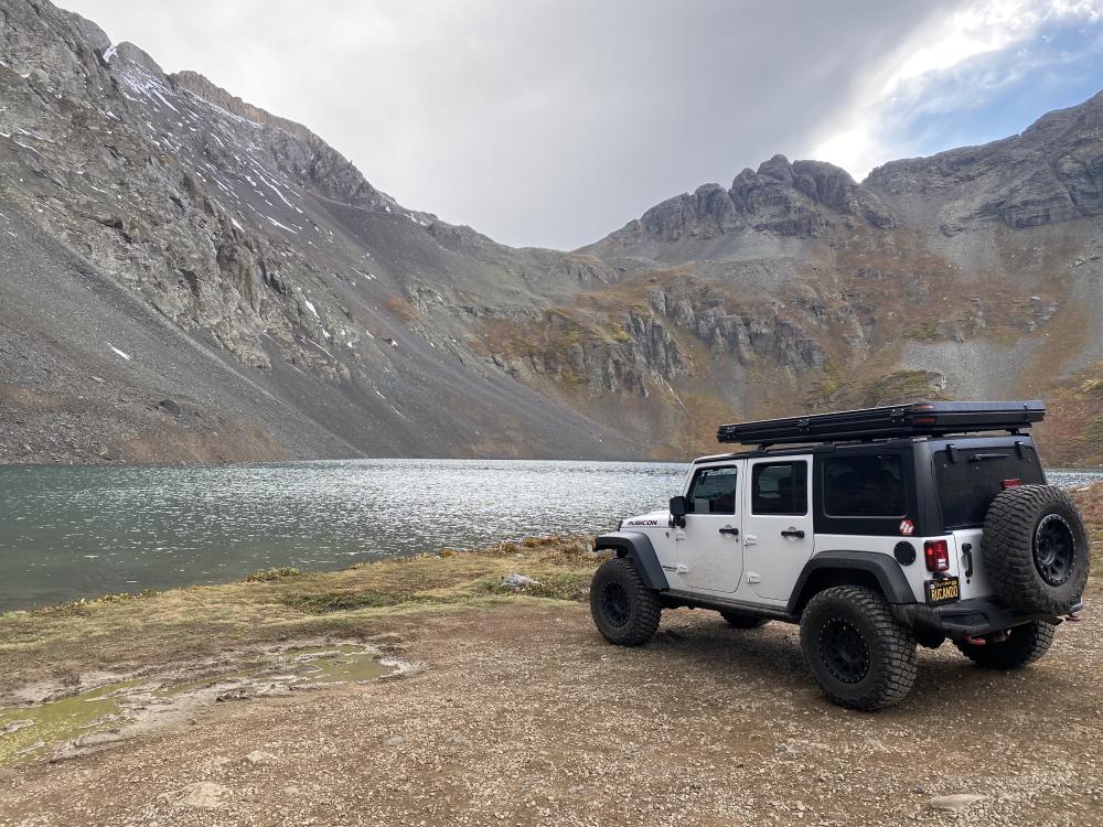 White Jeep Wrangler parked beside an alpine lake near Silverton, framed by steep, snow-dusted mountains under a stormy sky.