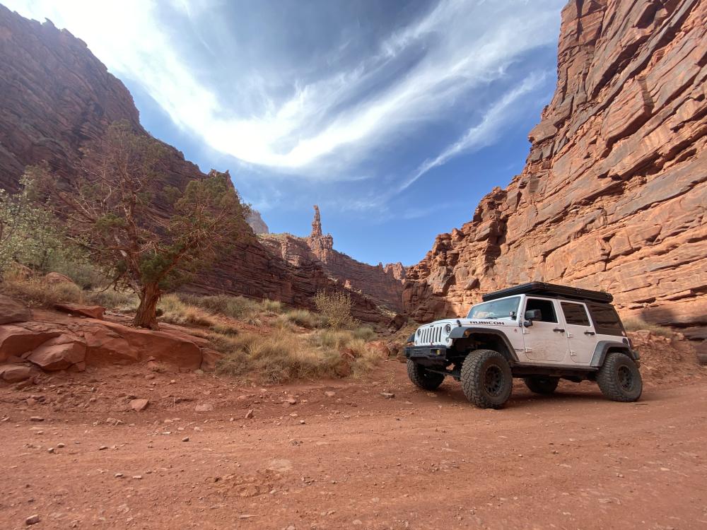 White Jeep Wrangler parked in a red rock canyon near Moab, with a slender sandstone spire rising in the distance.