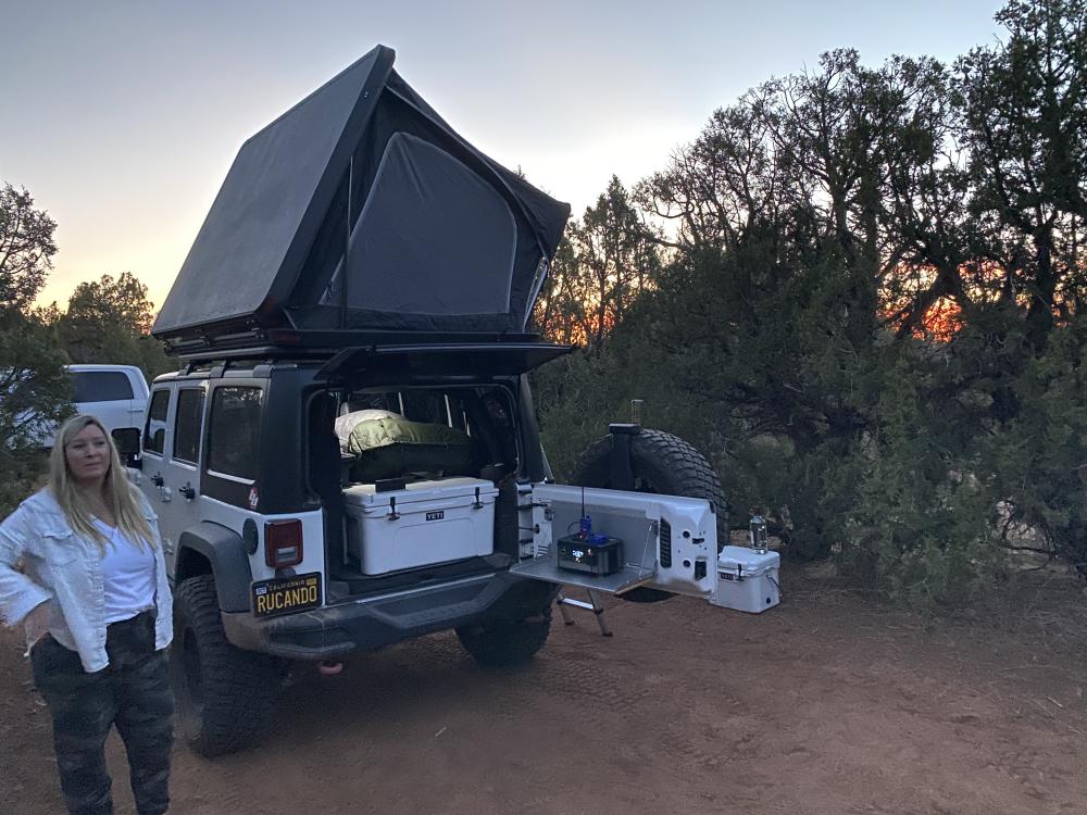 Woman standing beside a Jeep Wrangler with its rooftop tent open at a dusty campsite above Lake Powell at sunset.