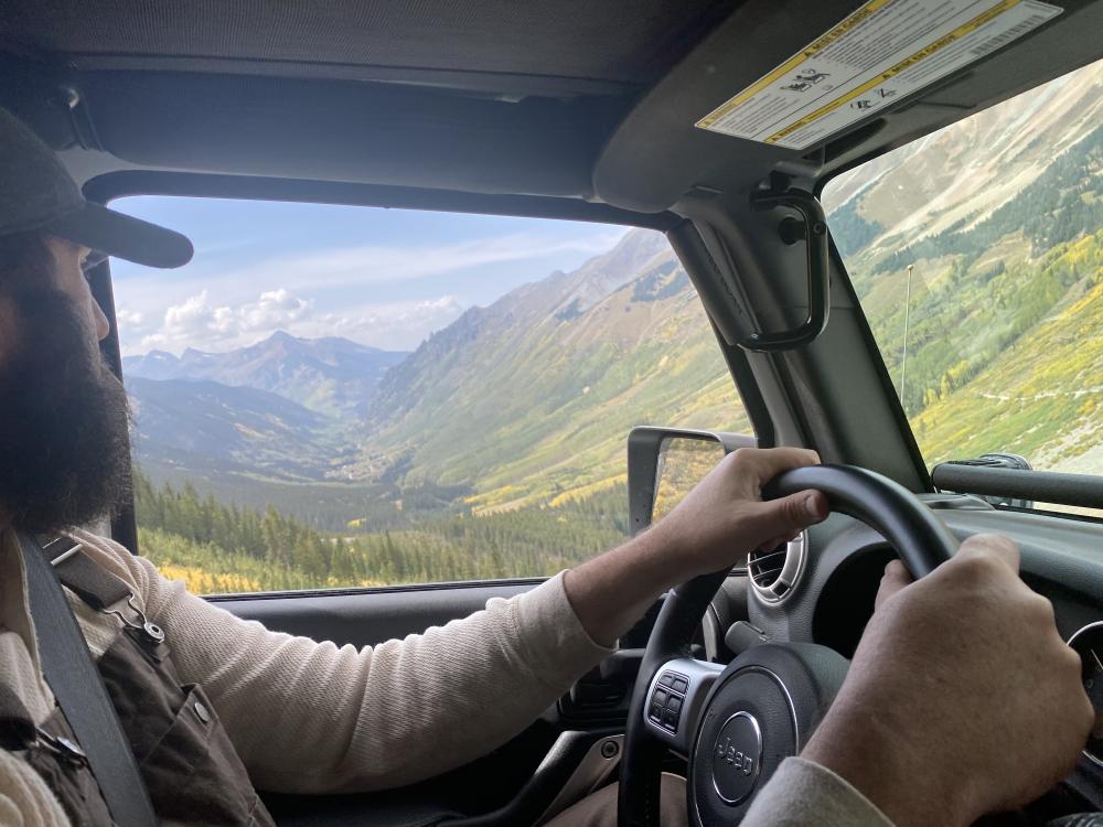 View from inside a Jeep of a driver's hand on the wheel, with a forested mountain valley visible through the windshield.