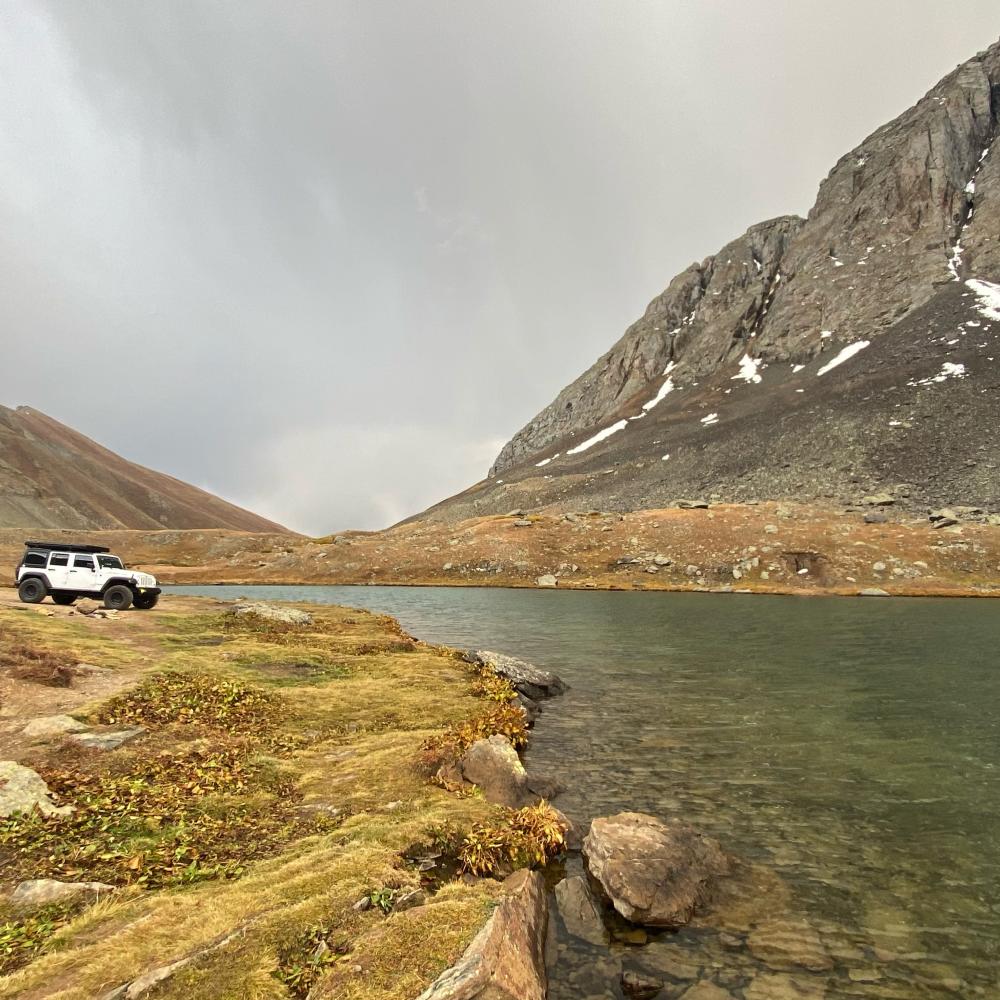 White Jeep Wrangler parked beside a turquoise alpine lake under stormy skies, with snow-streaked peaks rising behind it.