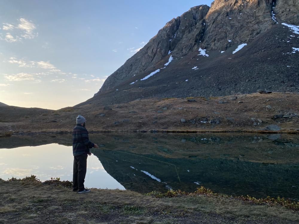 Man fly fishing at the edge of a still alpine lake at dusk, mountains reflected in the glassy water.