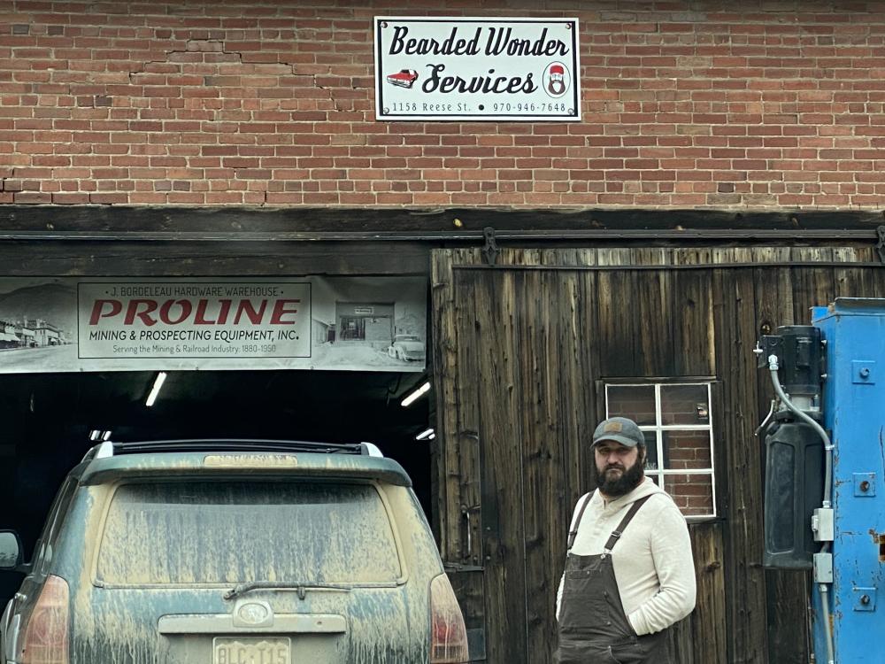 Man standing outside a rustic Silverton garage with a humorous 'Bearded Wonder Services' sign above the door.