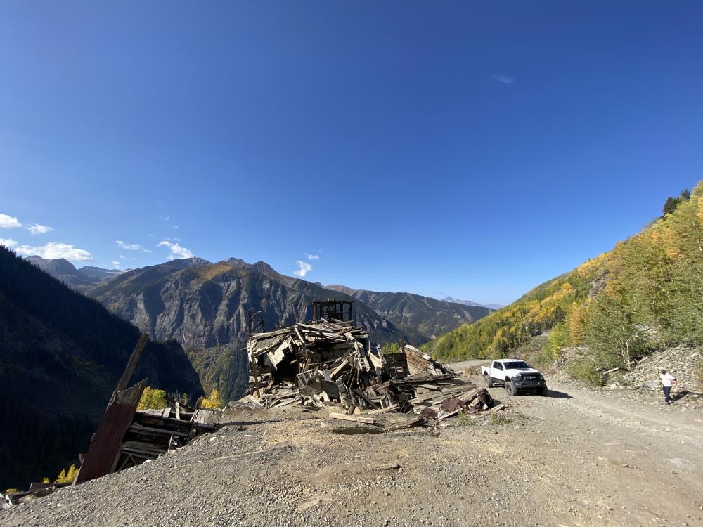 White pickup truck parked beside the collapsed wooden ruins of a mountainside mine building overlooking a Telluride valley.
