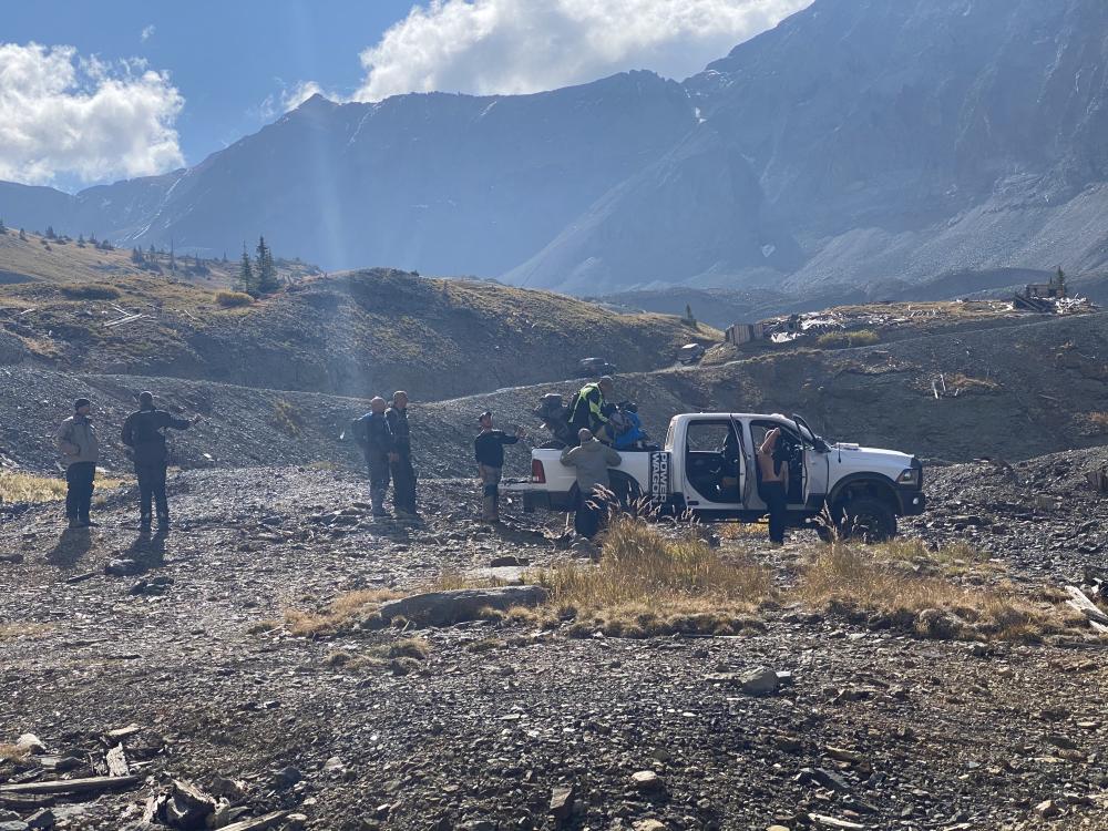 Group of people gathered around a white Ram Power Wagon at a high-altitude mining ruins site surrounded by rocky peaks.