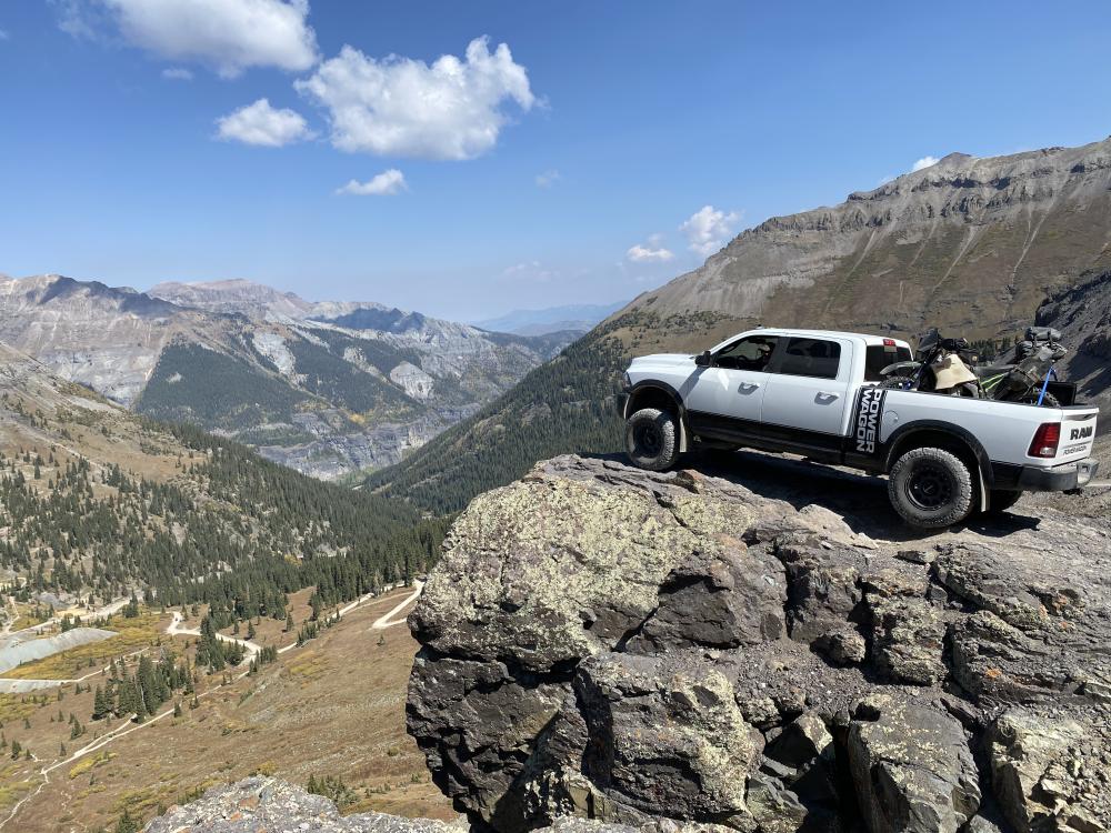White Ram Power Wagon with dirt bikes loaded in the bed, parked on a rock outcropping overlooking a forested mountain valley.