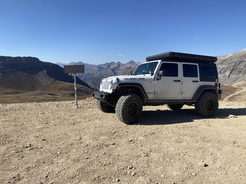 White Jeep Wrangler parked next to the Black Bear Pass summit sign marking 12,840 feet, with layered mountains behind it.