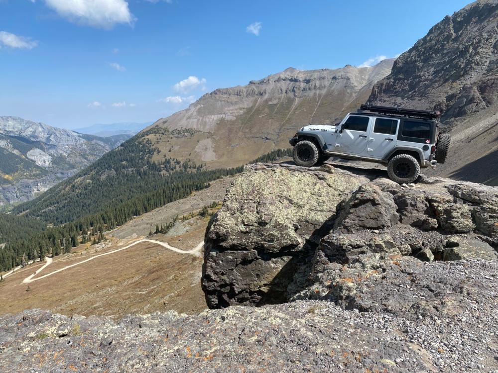 Silver Jeep Wrangler perched on a rocky outcropping high above a forested valley and winding dirt road.