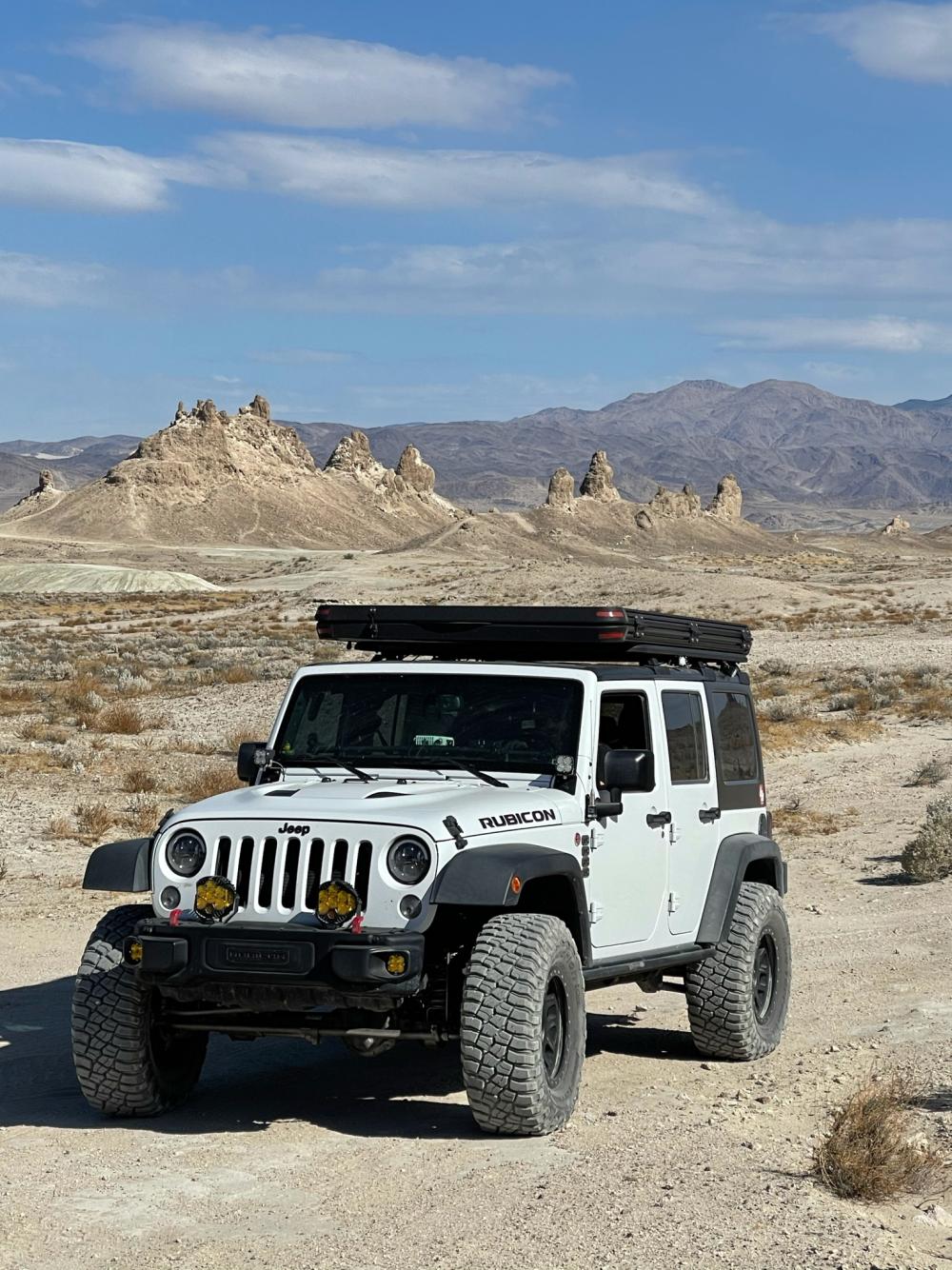 White Jeep Wrangler with a rooftop tent parked on a dirt road, with the Trona Pinnacles rock spires and mountains behind it.