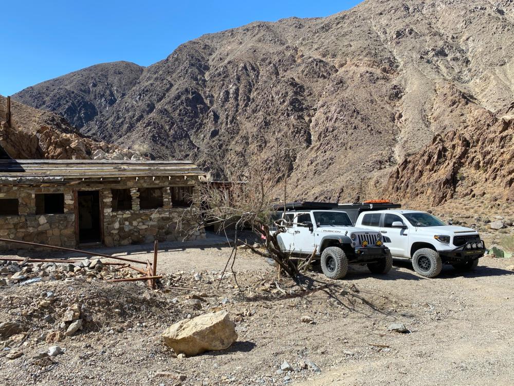 White Jeep Wrangler and Toyota Tundra parked beside an abandoned stone cabin, tucked against steep desert mountains.