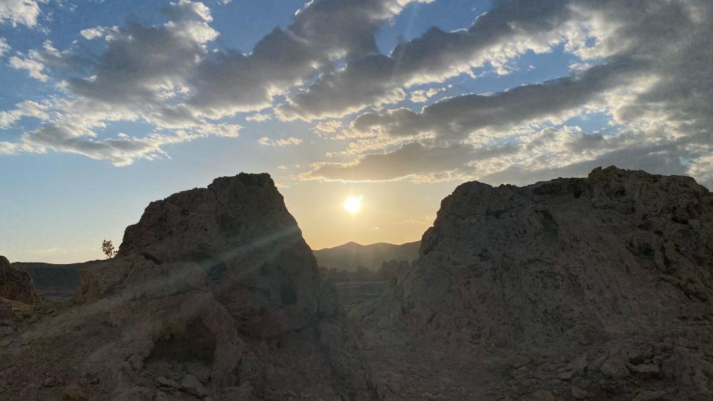 Sun setting between two rock formations in Death Valley, with distant mountains silhouetted against a cloud-streaked sky.