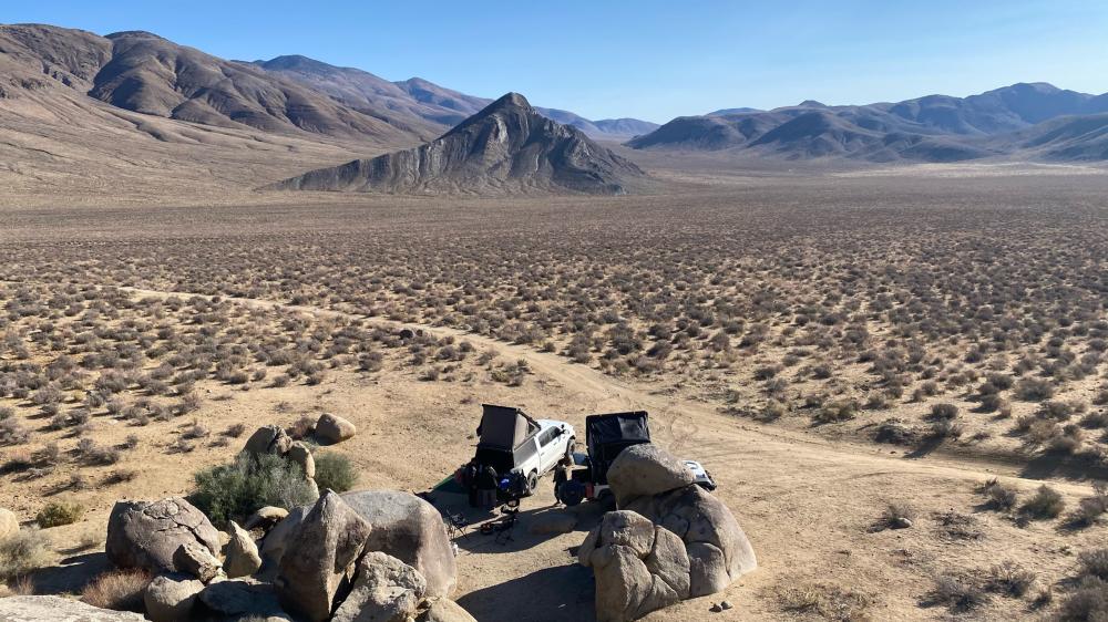Aerial view of a rooftop-tent campsite tucked among boulders overlooking a vast, empty desert valley ringed by mountains.