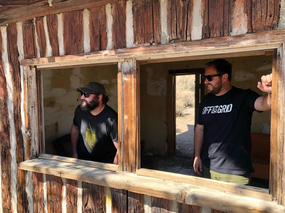 Two men looking out through the glassless window frames of an abandoned wooden mining cabin.