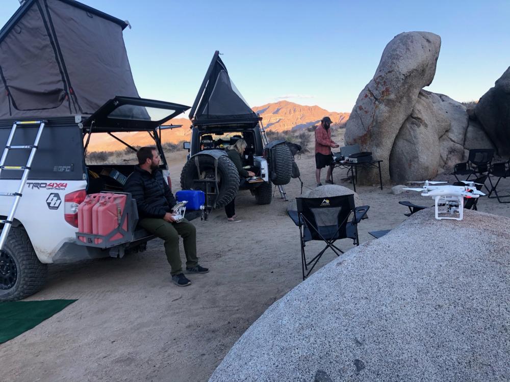 Group camped among boulders at dusk with rooftop tents open, a drone resting nearby ready for a flight.