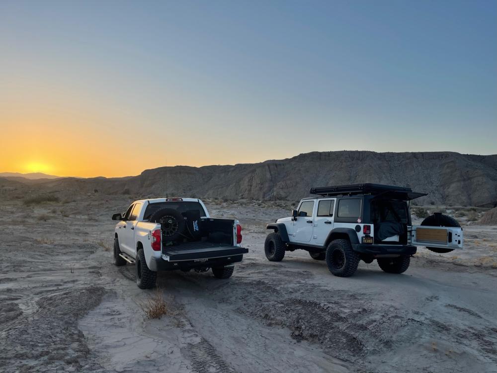 White Toyota Tundra and Jeep Wrangler parked in the wash at sunset, with the sun dropping behind distant badlands.