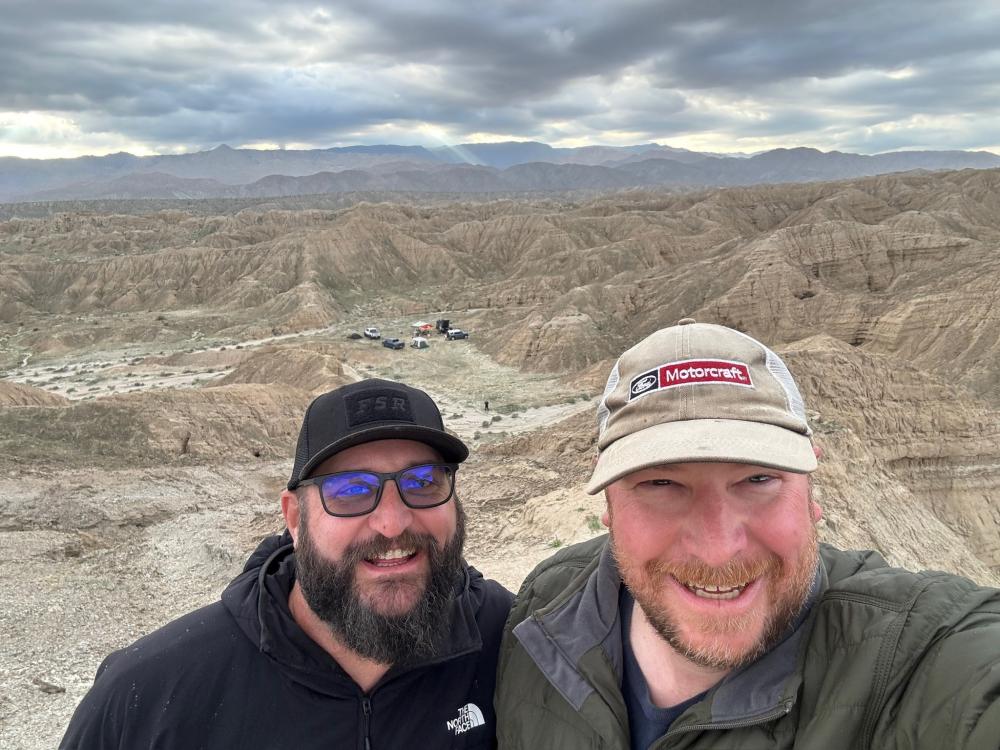 Two friends taking a selfie on a ridge above Anza-Borrego badlands, with a desert campsite visible far below.
