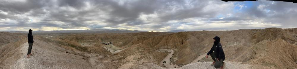 Wide panoramic view of two people standing on a badlands ridge overlooking a winding wash and distant camp, under a dramatic cloudy sky.