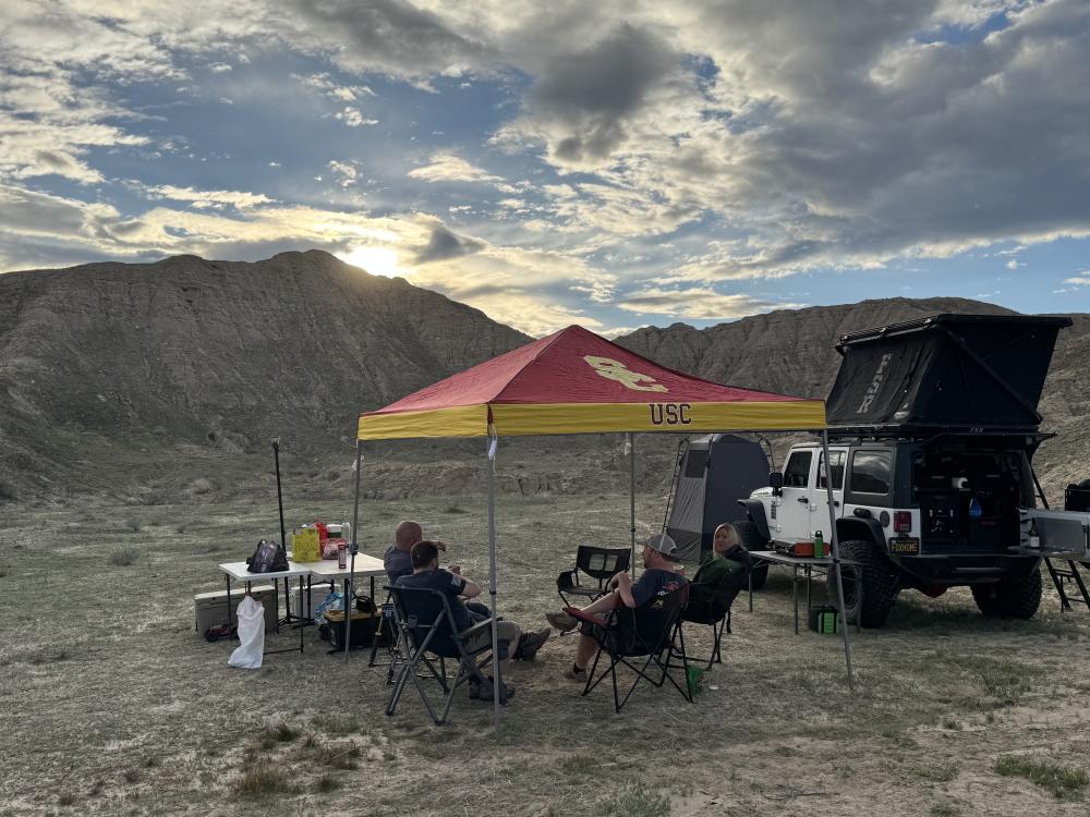 Group relaxing in camp chairs under a maroon and gold USC canopy at sunset, with a Jeep Wrangler and rooftop tent parked nearby.