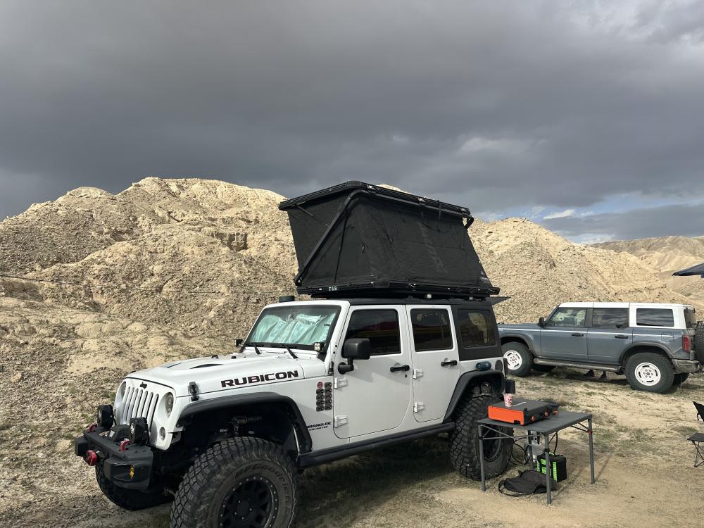 White Jeep Wrangler Rubicon with its rooftop tent popped up, parked on a dirt patch beneath a dark, storm-heavy sky.