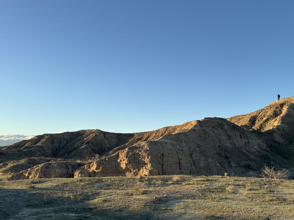 A lone silhouette standing atop a sunlit desert ridge under a clear blue sky, with rolling badlands below.