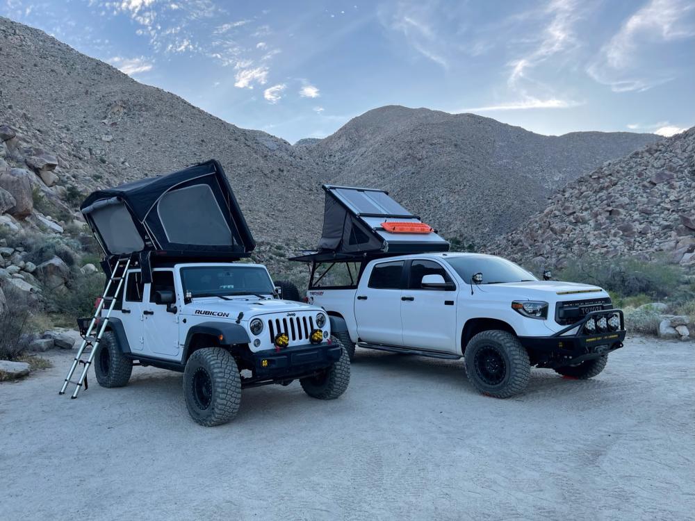 Jeep Wrangler and Toyota Tundra with rooftop tents open at camp, framed by boulder-strewn hillsides at dusk.