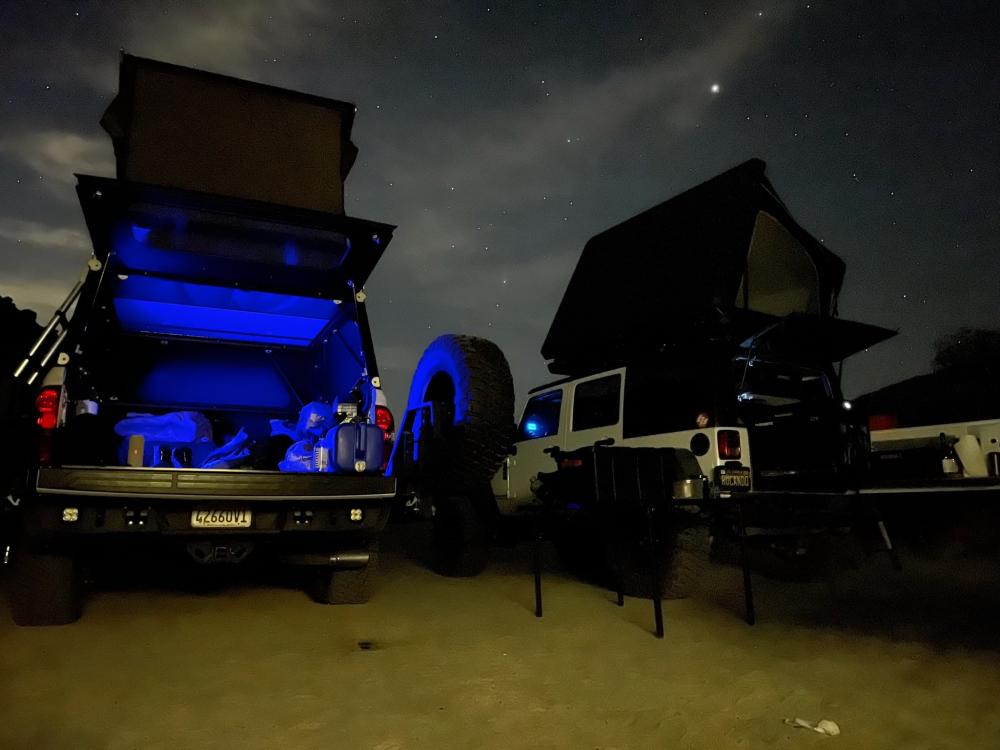 Truck bed lit with blue LED lighting at night, parked alongside a Jeep Wrangler at a starlit desert campsite.