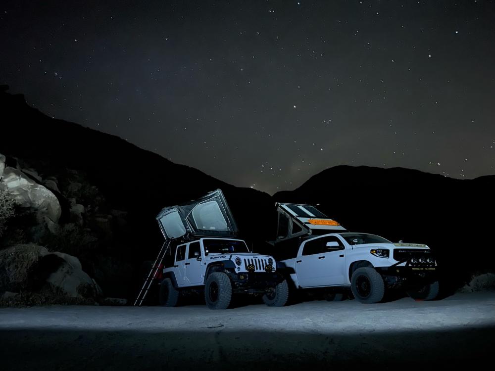 Jeep Wrangler and Toyota Tundra with rooftop tents open, illuminated by headlights beneath a star-filled night sky.
