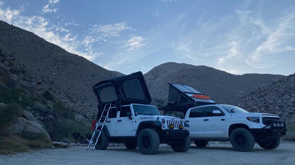 Jeep Wrangler and Toyota Tundra with rooftop tents open at a boulder-lined desert campsite at dawn, with camp chairs set up nearby.