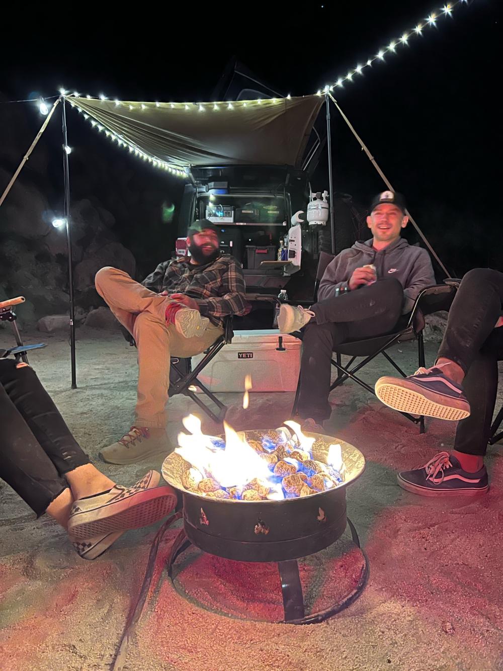 Group relaxing around a propane fire pit at a nighttime desert camp, with string lights strung overhead.
