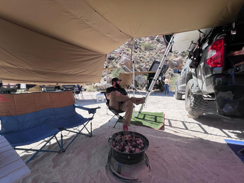 Man relaxing in a camp chair beneath a large shade canopy at a rocky desert campsite, with rigs and gear parked nearby.