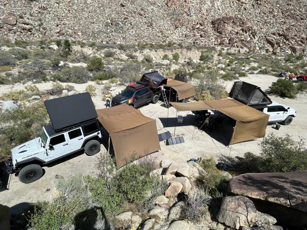 Aerial view of a multi-vehicle desert campsite with several shade canopies set up between Jeeps and trucks in a boulder-strewn canyon.