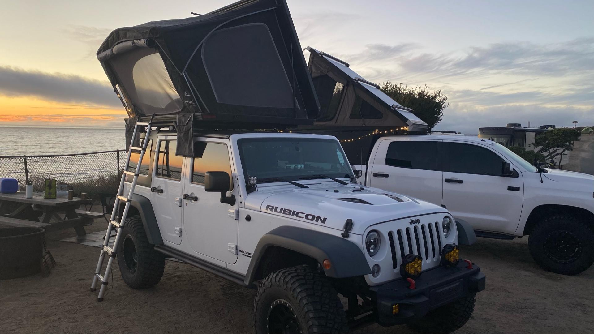 White Jeep Wrangler with its rooftop tent open at a bluff-top campground overlooking the ocean at sunset.