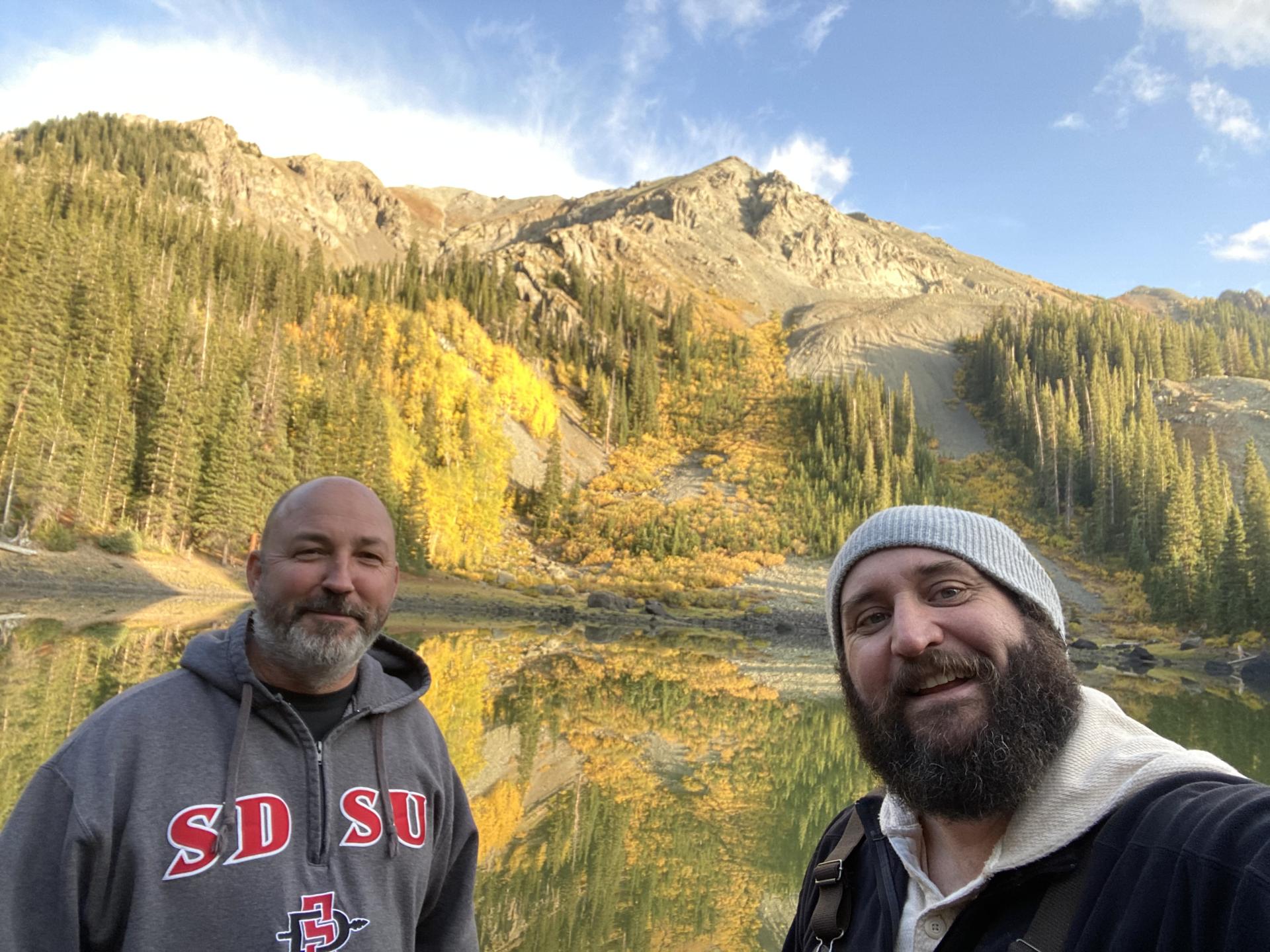 Shawn and a friend taking a selfie beside an alpine lake, with mountains and golden aspen trees reflected in the still water.