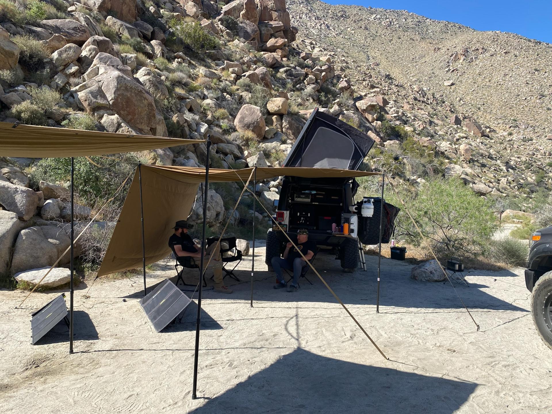 Two people relaxing in camp chairs under a shade canopy strung between a Jeep and a truck, with solar panels set up at a rocky desert campsite.